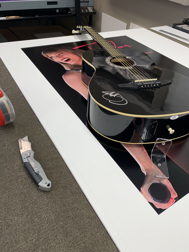 An image capturing the behind-the-scenes process of framing the signed guitar for the Big Stars & Pink Guitars fundraiser event. A black acoustic guitar, signed by Taylor Swift, is carefully positioned within a custom framing workshop. A skilled artisan from Piper Arts & Custom Framing is meticulously arranging the guitar within a stylish frame, ensuring that every detail is perfectly aligned. Tools and materials for framing are visible in the background, highlighting the craftsmanship and attention to detail involved in the framing process. This image offers a glimpse into the creation of the iconic piece of memorabilia that contributed to the success of the fundraising event.