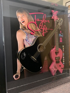 An image of a black acoustic guitar mounted in a custom frame. The frame features a pink FireFly guitar alongside an authenticated autograph by Taylor Swift. The background showcases the vibrant atmosphere of the Big Stars & Pink Guitars fundraiser event, with attendees gathered around to admire the stunning piece of memorabilia. The frame is elegantly displayed, capturing the essence of the event's record-breaking achievement.