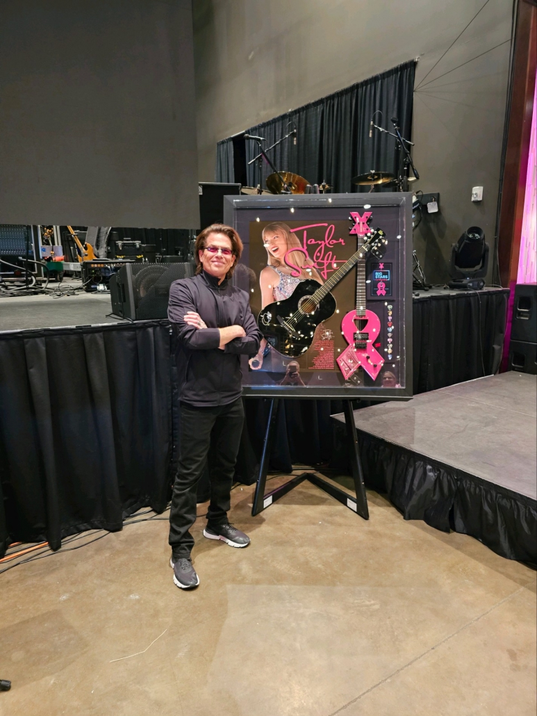 An image of Kory Van Sickle, the creator of Firefly Guitars, standing proudly next to a custom-framed black acoustic guitar. The guitar is mounted in a stylish frame and features a pink FireFly guitar alongside an authenticated autograph by Taylor Swift. Kory is smiling and gesturing towards the guitar, showcasing the collaborative masterpiece created for the Big Stars & Pink Guitars fundraiser event. The background captures the excitement and energy of the event venue, with attendees admiring the impressive display.