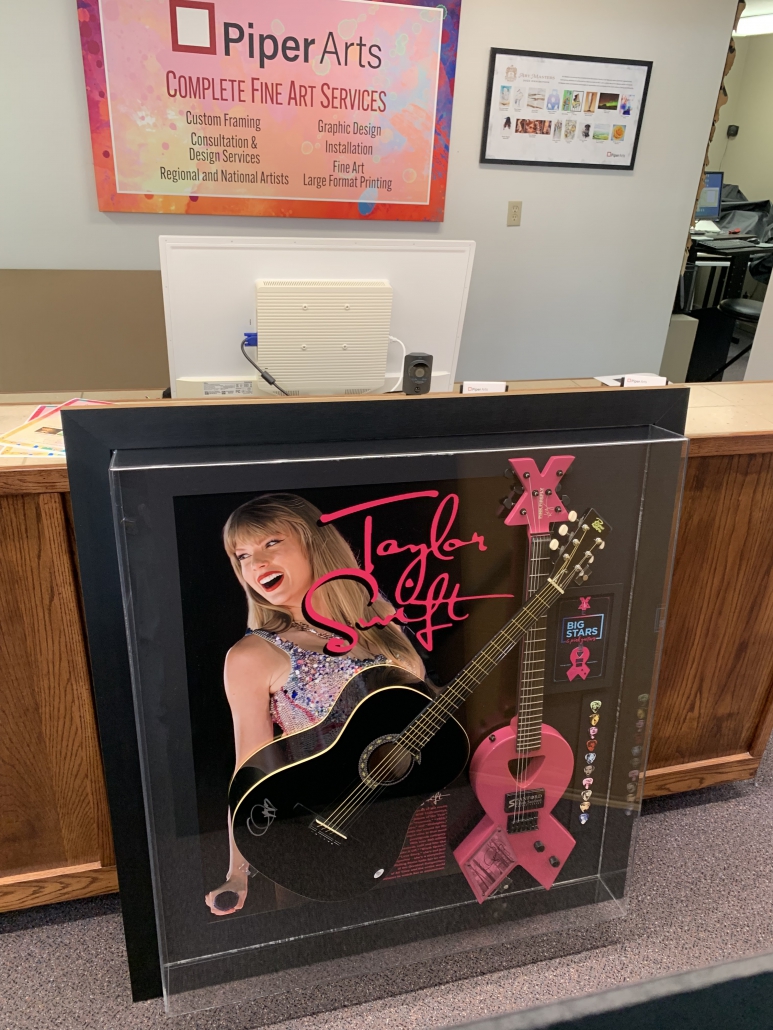 An image showcasing the finished framed piece displayed in the gallery at Piper Arts. The custom-framed black acoustic guitar, signed by Taylor Swift and accompanied by a pink FireFly guitar, is elegantly mounted within a stylish frame. The gallery setting provides a serene backdrop, with soft lighting illuminating the framed artwork and emphasizing its beauty and craftsmanship. The framed piece is meticulously presented, reflecting the care and attention to detail that went into its creation. This image captures the anticipation and excitement surrounding the masterpiece before it was showcased at the Big Stars & Pink Guitars fundraiser event.