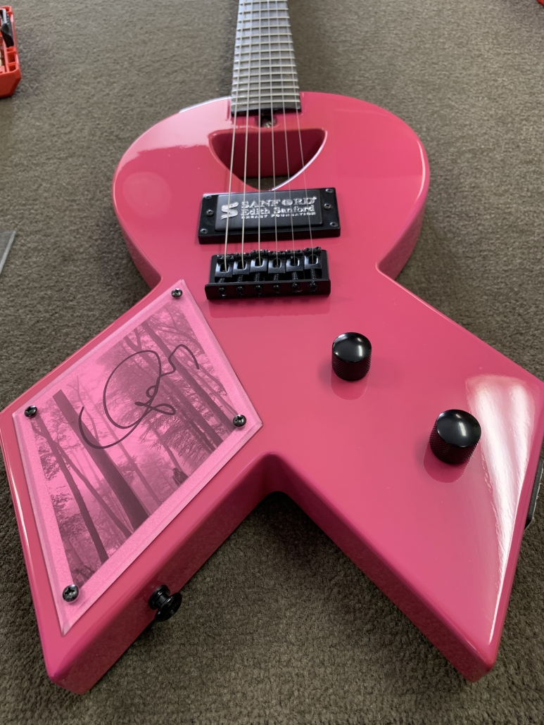 An image of a stunning pink guitar, crafted to resemble a ribbon for breast cancer awareness. The guitar features intricate details and vibrant hues, symbolizing hope, strength, and support for those affected by breast cancer. This iconic Pink Guitar is a signature piece created by Kory Van Sickle of Firefly Guitars, and it serves as the centerpiece of the annual Big Stars & Pink Guitars fundraiser event. The guitar is elegantly displayed, showcasing its beauty and significance in raising awareness and funds for breast cancer research.